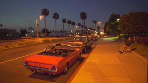 1995 Ford Taurus - A red 1965 Ford Mustang convertible is parked on the side of the road as dusk settles, with palm trees lining the street.
