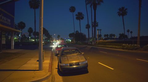 1995 Ford Taurus - A dark sedan is parked on the side of a road at night, with palm trees lining the street.