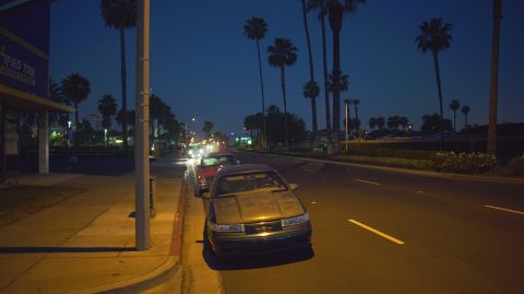 1995 Ford Taurus - Multiple vehicles are parked along a street at night, with palm trees lining the background.
