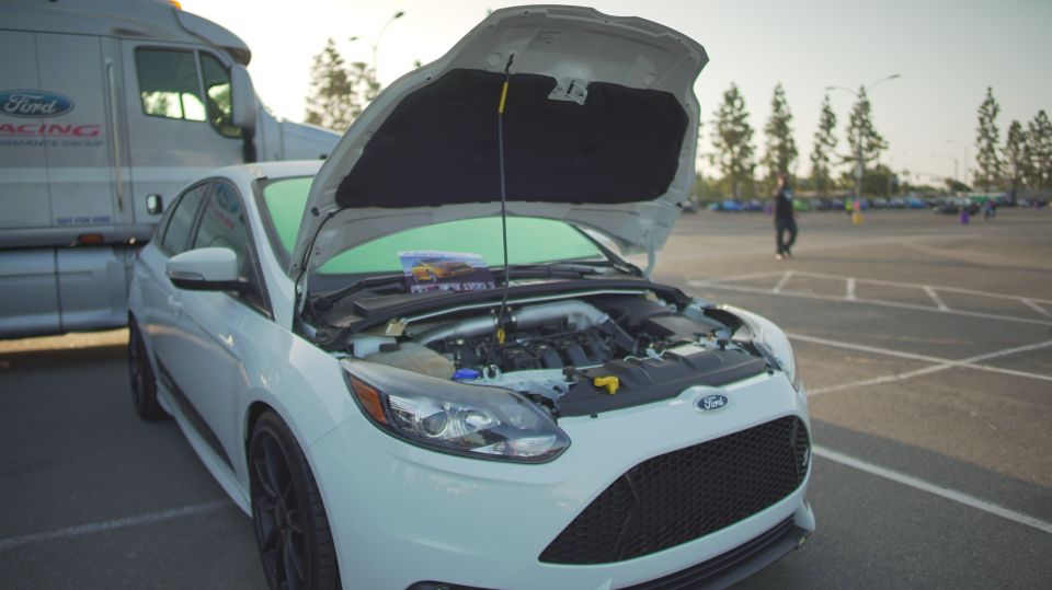 White Ford Focus ST, front view, hood open, showing engine. A Ford Racing truck is parked behind it