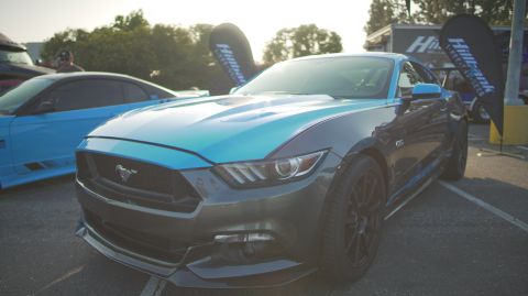 1995 Ford Taurus - A modern Ford Mustang GT is parked at an outdoor event with other vehicles and banners.