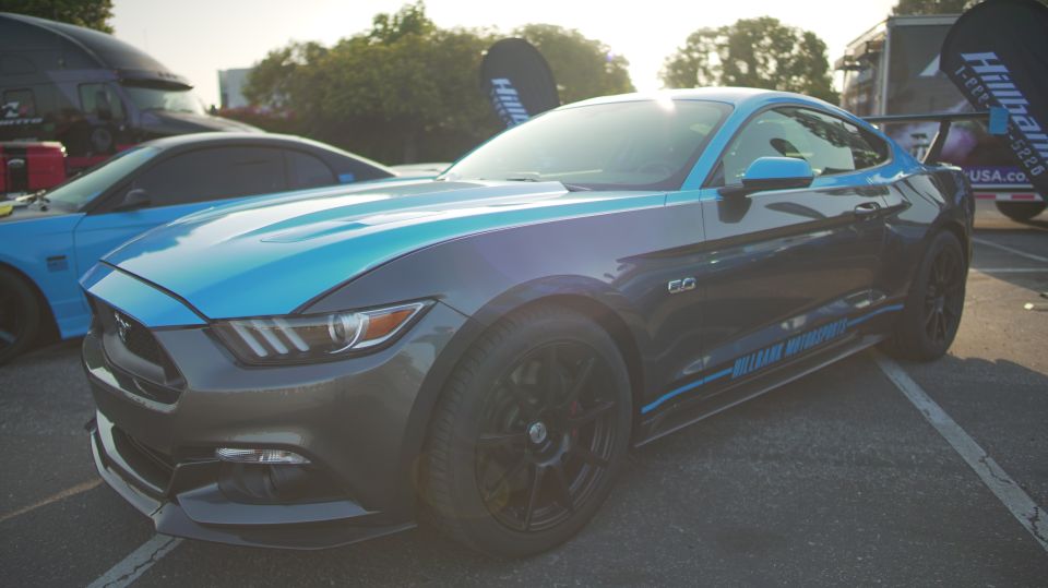 Gray and blue Ford Mustang with racing stripes and a rear spoiler. Text on door reads 'BILLY'S MOTORSPORTS'. Another blue car is visible behind it