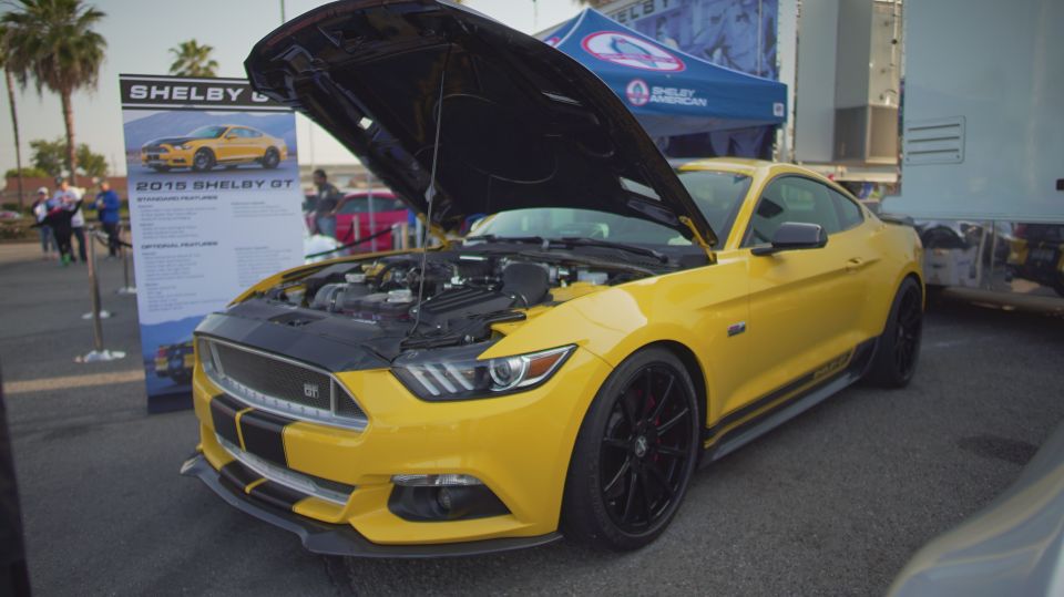 Yellow 2015 Shelby GT Mustang with black racing stripes, hood open showing engine. Display sign behind it
