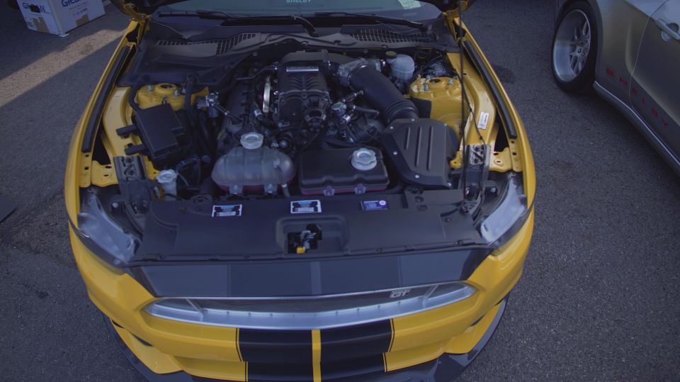Yellow Ford Mustang with hood open showing engine and supercharger. Another car is parked to the right