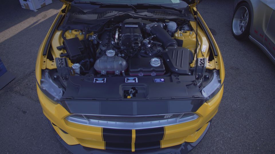 Yellow Ford Mustang with hood open showing engine and supercharger. Another car is parked to the right