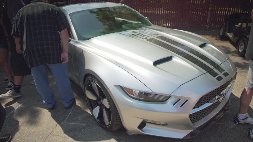 Silver Ford Mustang with black racing stripes and '725' on the hood, viewed from the front-right