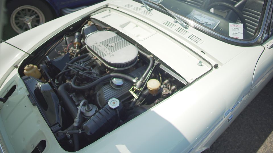 Engine bay of a white car, with another car's wheel visible in the background. A 'For Sale' sign is on the windshield
