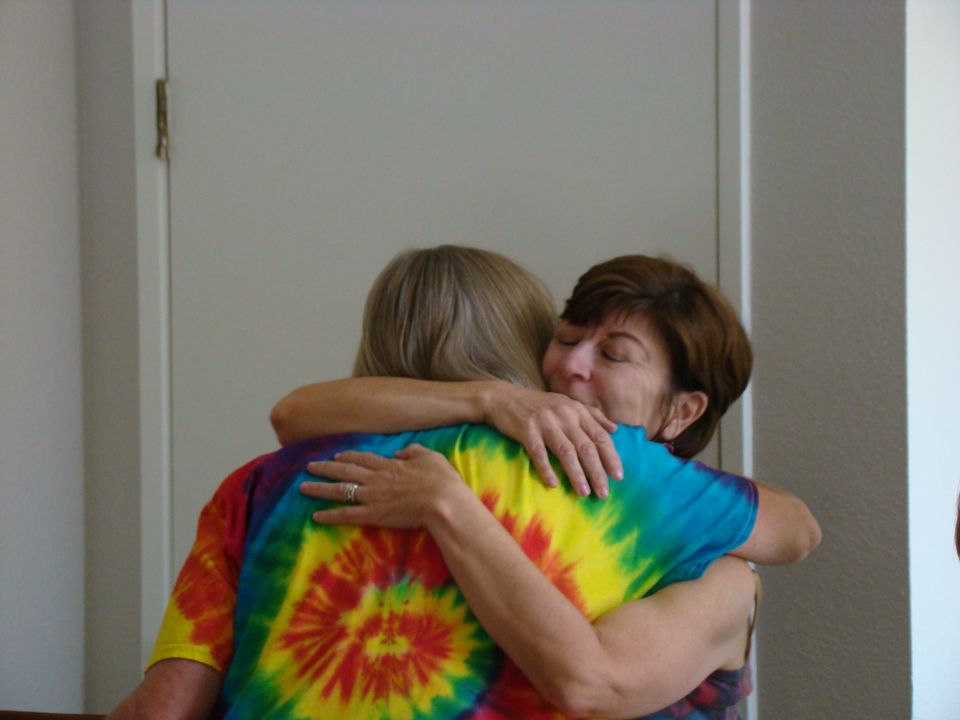 Two women embrace, one wearing a tie-dye shirt. A closed door is behind them
