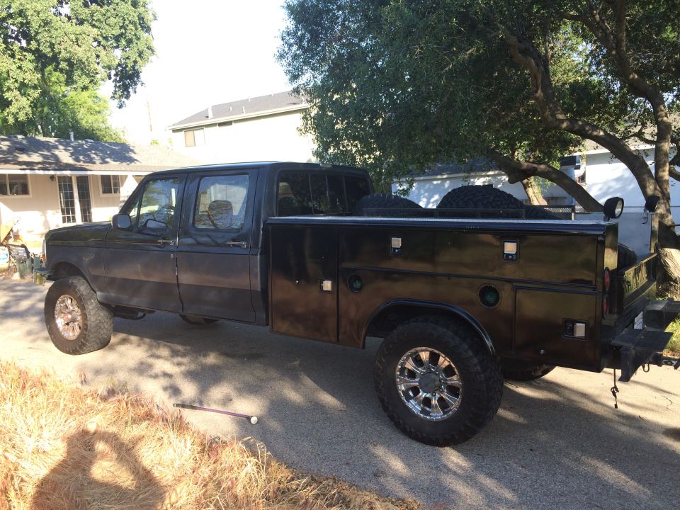 Black crew cab pickup truck with utility bed, chrome wheels, and spare tires in the bed. Visible from the side