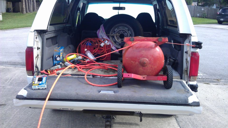 Red air compressor, hoses, and tools loaded in the bed of a white pickup truck. Spare tire visible in cab