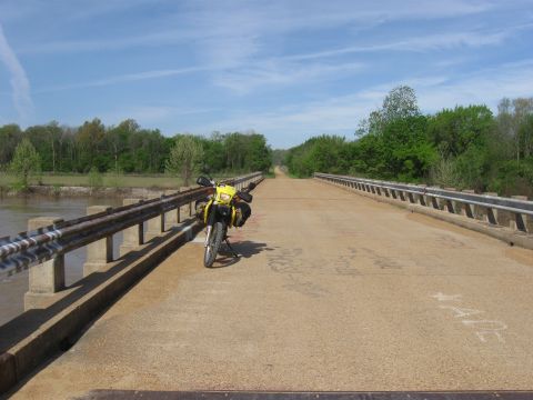 2006 Suzuki DR-Z400S - A lone yellow motorcycle rests on a bridge spanning a river, surrounded by trees under a blue sky.