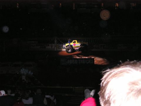 1996 Ford Bronco - A monster truck with a yellow body and diamond decal is spotlighted on a dirt arena floor.