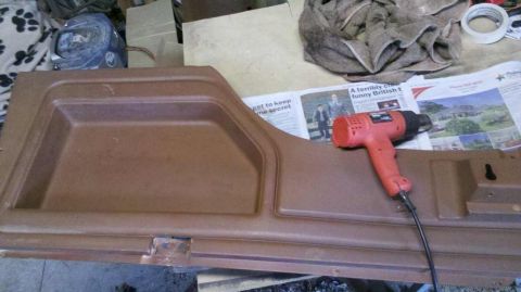 1982 Ford Bronco - A heat gun rests on a newspaper next to a brown car door panel in a workshop.