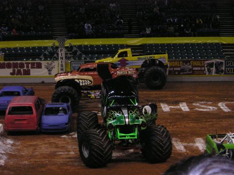 1996 Ford Bronco - Monster trucks are lined up on a dirt arena with crushed cars as obstacles.