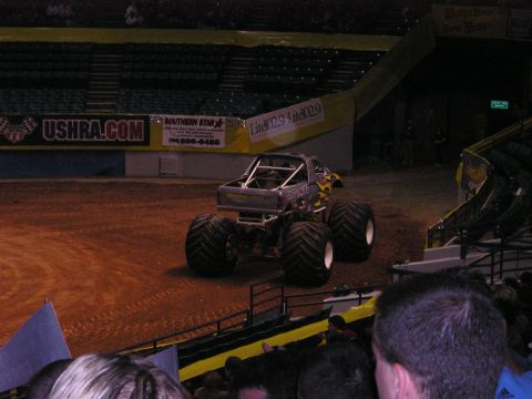 1996 Ford Bronco - A monster truck with flame decals sits on a dirt arena floor, viewed from the rear.
