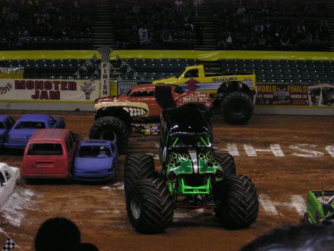 1996 Ford Bronco - Monster trucks and crushed cars are staged on a dirt arena, with a "Monster Jam" banner in the background.