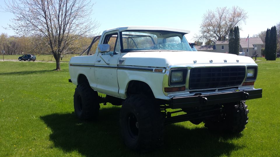 White lifted Ford Bronco with large tires parked on grass. A black car is visible on a road in the background