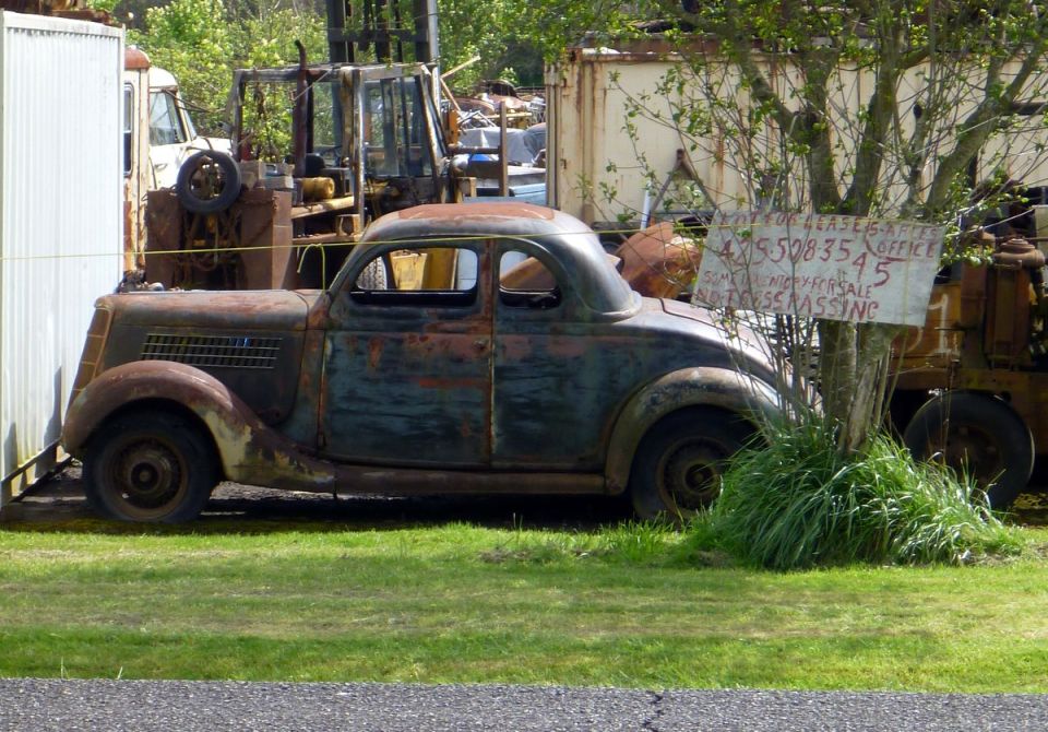 Rusty coupe with a grille resembling a 1936 Ford, seen from the side in a junkyard
