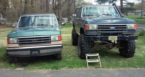 1990 Ford Bronco - A stock green Ford Bronco sits next to a heavily modified dark green Ford Bronco with large tires and a snorkel.