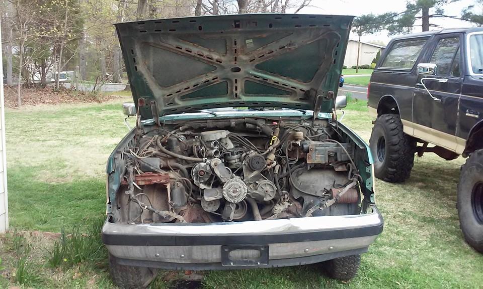 Green truck engine bay with hood open; black Bronco in background