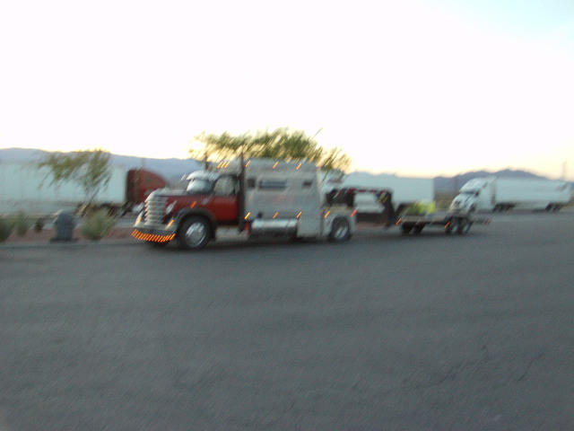Tow truck with a flatbed trailer carrying a small vehicle, parked near other semi-trucks