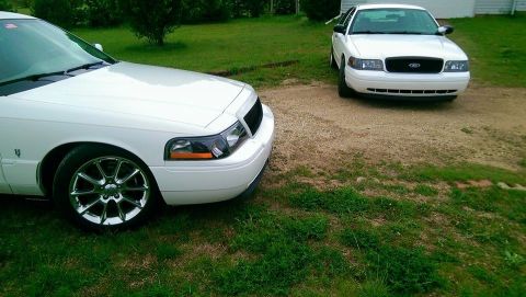 2000 Mercury Grand Marquis - Two white sedans, one a 2000 Mercury Grand Marquis, are parked on a gravel and grass area.
