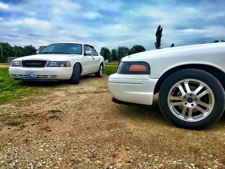 Two white sedans parked on a gravel road, one in the foreground showing its front wheel and fender, the other in the background