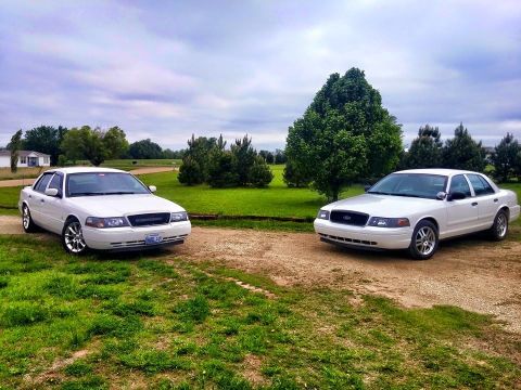 2000 Mercury Grand Marquis - Two white 2000 Mercury Grand Marquis sedans are parked side-by-side on a dirt road with a grassy field and trees in the background.