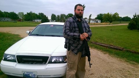 2000 Mercury Grand Marquis - A man stands with a rifle next to a white police car on a dirt road.
