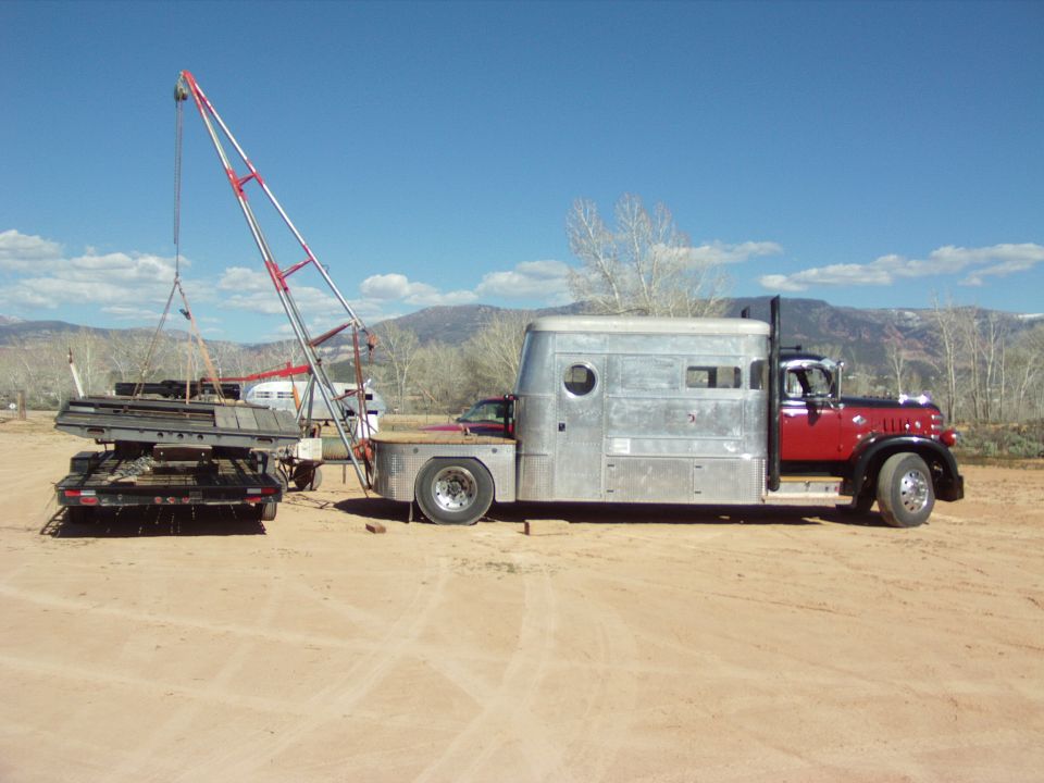 Truck with silver trailer body and red cab, crane lifting metal onto a flatbed trailer