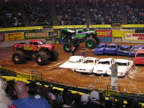 1996 Ford Bronco - Monster trucks perform in an arena, crushing a line of cars as part of the show.