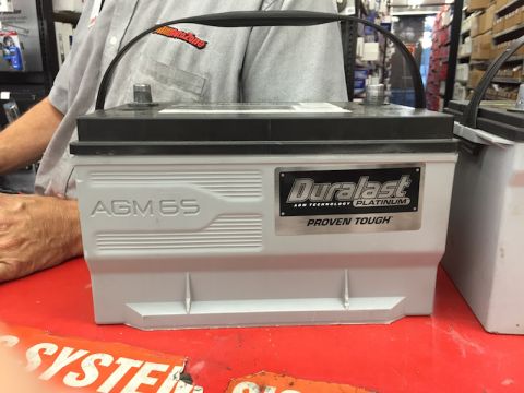 1989 Ford Bronco - A Duralast AGM battery is displayed on a red surface, with a person in an AutoZone shirt in the background.