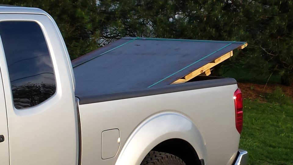 Silver pickup truck bed with a dark tarp over wooden supports, seen from the rear quarter