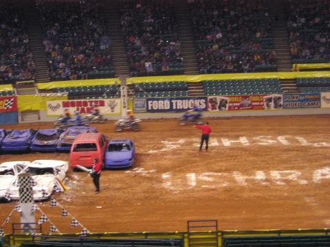 1996 Ford Bronco - A monster truck event is underway in an arena, with several crushed cars on the dirt track and spectators in the stands.