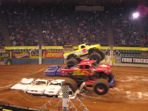 1996 Ford Bronco - Two monster trucks perform stunts over a pile of crushed cars in front of a large crowd.