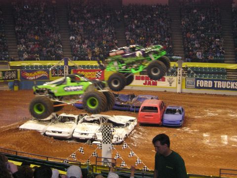 1996 Ford Bronco - Monster trucks soar over a pile of wrecked cars in an arena, thrilling the crowd.