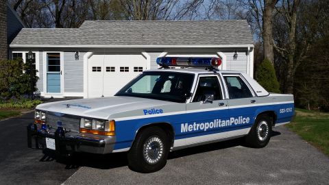 1991 Ford LTD Crown Victoria - A police car with 'Metropolitan Police' text and emergency lights is parked in front of a suburban garage.