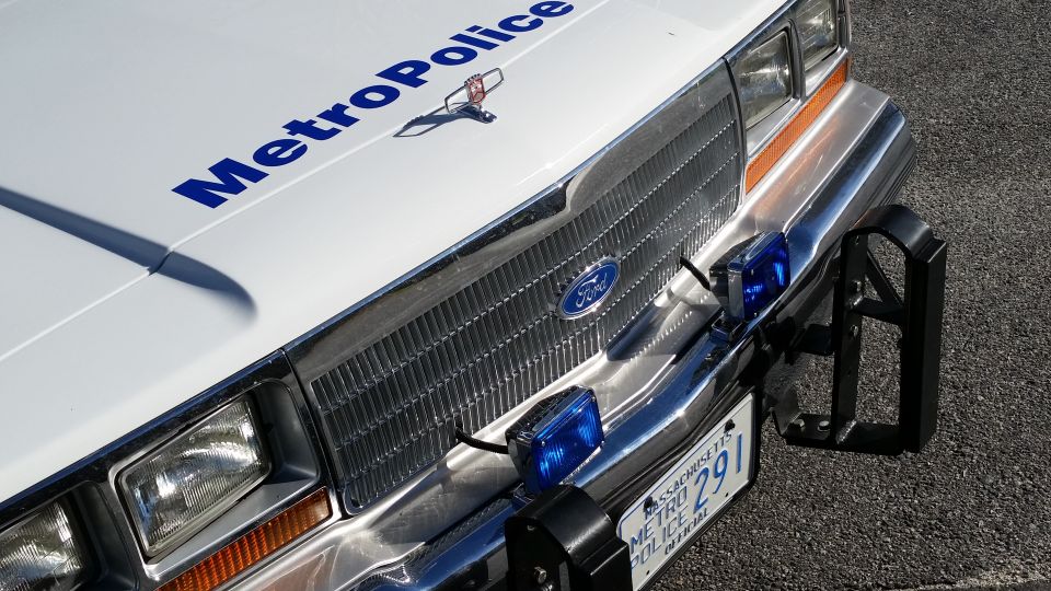 Front of a white police car with 'Metro Police' on hood, Ford emblem on grille, and blue lights
