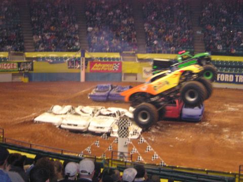 1996 Ford Bronco - Monster trucks perform stunts in front of a crowd in a stadium.