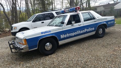 1991 Ford LTD Crown Victoria - Police car with 'Metropolitan Police' text and light bar parked on gravel next to an SUV.