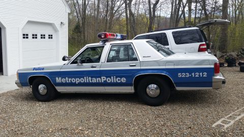 1991 Ford LTD Crown Victoria - A side view of a police car with a light bar on top, parked next to a garage with another vehicle behind it.