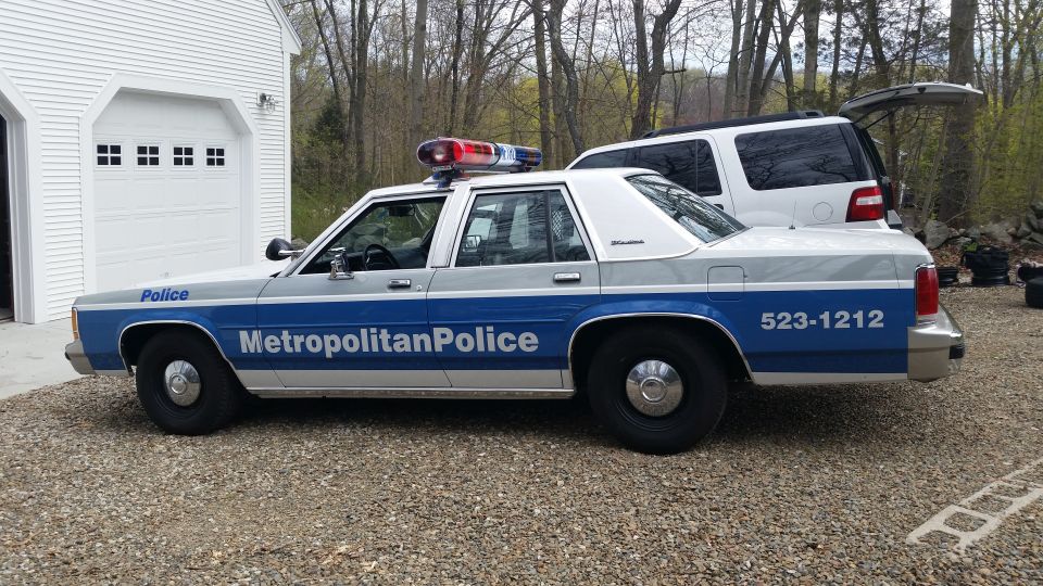 Police car with 'Metropolitan Police' and phone number 523-1212 on the side, parked in front of a garage with a white SUV behind it
