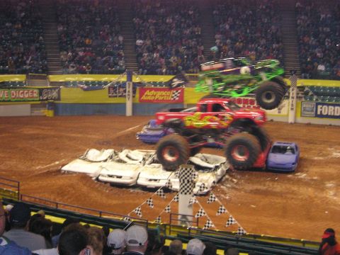 1996 Ford Bronco - Two monster trucks perform stunts, jumping over a pile of cars in an arena.