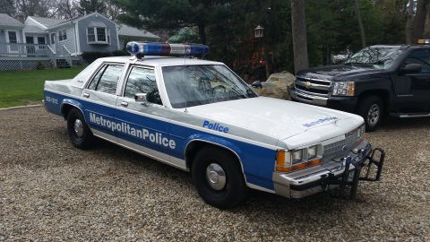 1991 Ford LTD Crown Victoria - A police car with a light bar on top is parked on a gravel driveway next to a house.