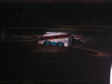 1996 Ford Bronco - A custom trailer, likely for a monster truck, sits on a dirt arena floor under stadium lights.