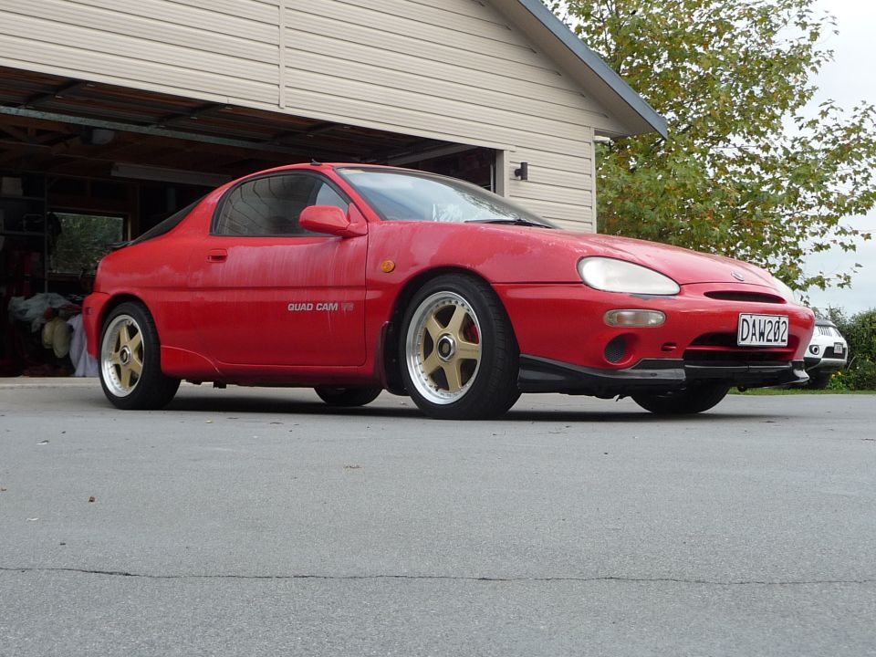 Red 1993 Mazda MX-3 coupe with gold five-spoke wheels parked in front of a garage