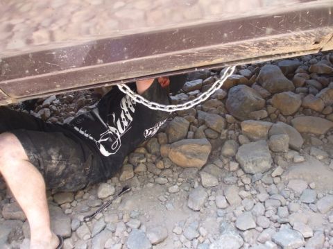 1979 Ford Bronco - A person is seen working on the underside of a vehicle, with a chain draped across the frame and a wrench on the ground.
