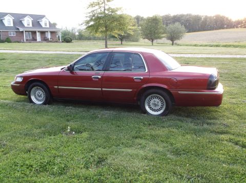1999 Mercury Grand Marquis - Red sedan with flat tires parked on grass in front of a house.