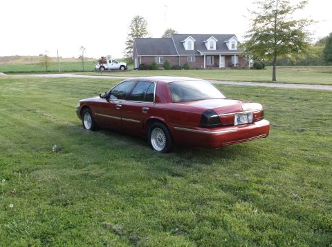 1999 Mercury Grand Marquis - A red 1999 Mercury Grand Marquis is parked on the grass in front of a house, with a white pickup truck visible in the distance.