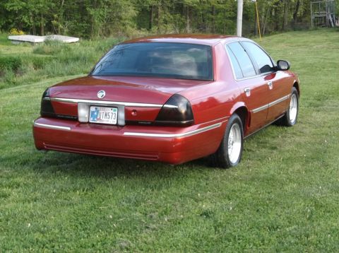 1999 Mercury Grand Marquis - Rear view of a red 1999 Mercury Grand Marquis parked on a grassy area with trees in the background.